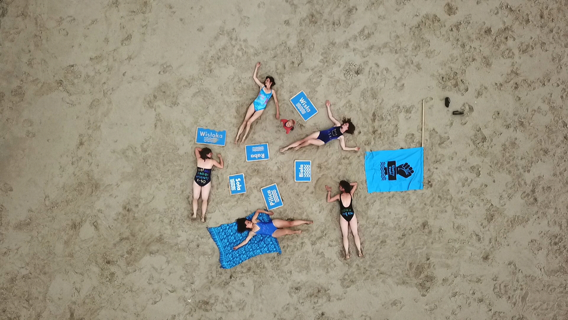 Zdjęcie przedstawia grupę kobiet leżących w kostiumach na plaży. Zdjęcie wykonane z lotu ptaka, od góry. Kobiety w kostiumach kąpielowych leżą na piasku, wokół porozrzucane niebieskie tabliczki z nazwami rzek Wisła, Raba , Biała, Wisłoka, Soła. Obok nich rzucona na piach niebieska flaga, z czarnym nadrukiem loga Siostry Rzeki, które przedstawi pięść zaciśniętą wynurzającą się z wody,. Wśród leżących kobiet stoi chłopiec patrzący w górę, widzimy jego wzrok.