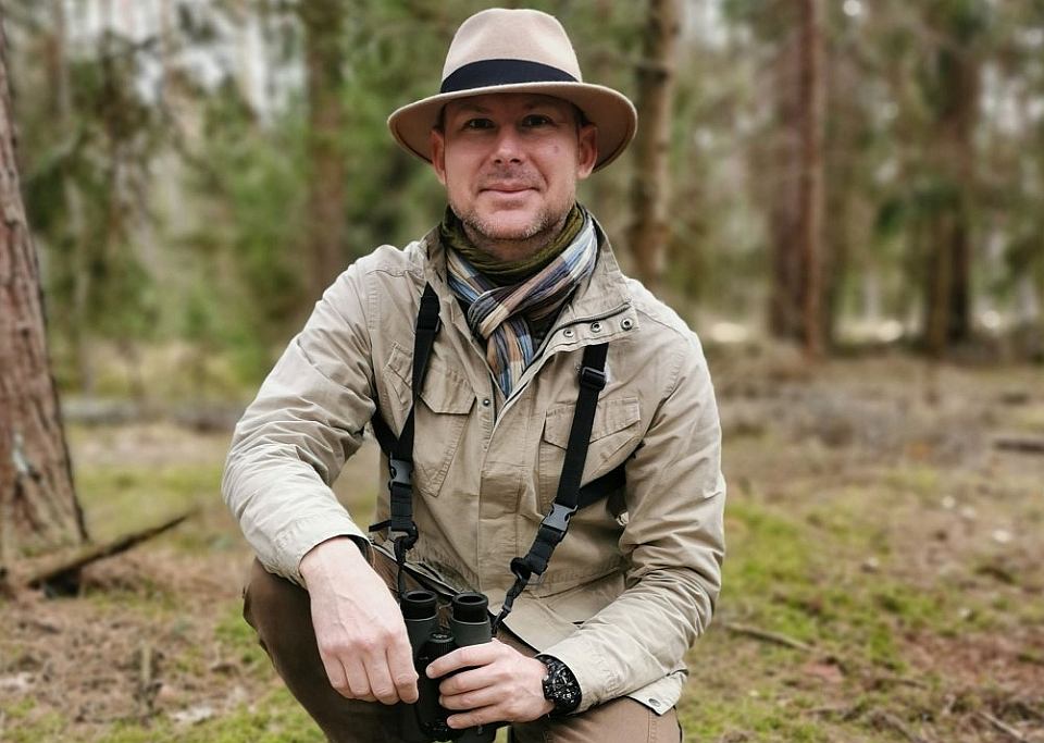 Na zdjęciu: Adam Zbyrt w lesie, ubrany w kapelusz, w dłoniach trzyma lornetkę. Przyklęka na jedno kolano. Fot. zasoby Adama Zbyrta